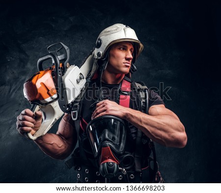 Muscular pensive man in helmet is looking away. He put his chainsaw on shoulder. With another hand he holds oxygen mask. Dark background.