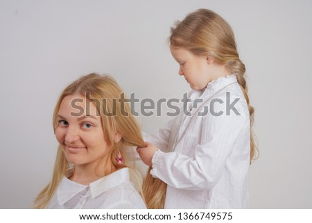 mom and daughter in white shirts with long blonde hair posing on a solid background in the Studio. a charming family takes care of each other and makes braids.