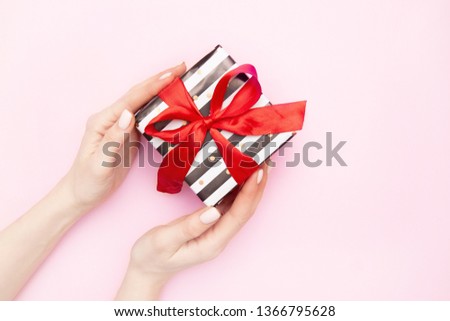 woman hands with Gift present box in white and black stripes with a red ribbon bow isolated on pink table top view. Minimal flat lay composition for sales, birthday, mother day or christmas.copyspace