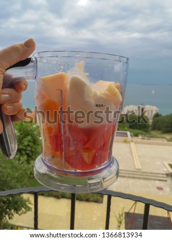 Blender bowl with large cut pieces of fruit and a few spoons of ice cream in a woman's hand against the blue sea and gray sky