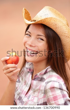 Peach eating cowgirl happy. Portrait of American cowgirl eating peach or nectarine fruit smiling and laughing wearing cowboy hat outside. Healthy eating concept with multiracial Caucasian Asian girl