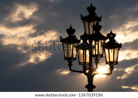 Old forging lamppost with five lanterns in Paris (France) and beautiful sunset with clouds and sun glow at background. Sunlight beams reflection.