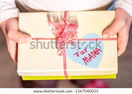 Little child holding gift box with ribbon and heart for her mum - Translation: for mum.