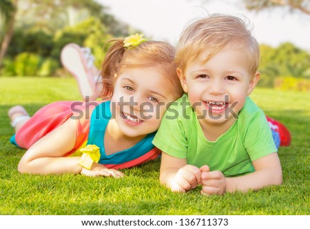 Picture of brother and sister having fun in the park, two cheerful children laying down on green grass, little girl and boy playing outdoors, best friends, happy family, love and happiness concept Royalty-Free Stock Photo #136711373