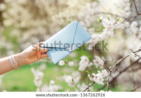 Close-up photo of female hands holding a blue invitation envelope with a wax seal, a gift certificate, a postcard, a wedding invitation card on the background of blooming flowers