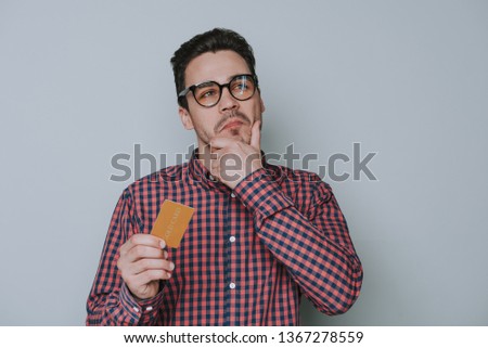 Waist up of pensive brown-haired man holding golden credit card isolated on gray background. He is wearing plaid shirt and looking away. He is touching his chin with arm in studio. Human emotions and