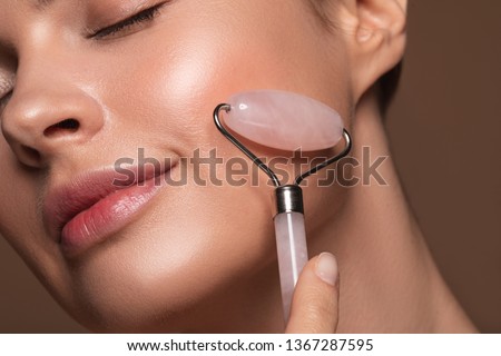 Close up photo of a young woman looking relaxed and smiling while using a natural rose quartz face roller Royalty-Free Stock Photo #1367287595