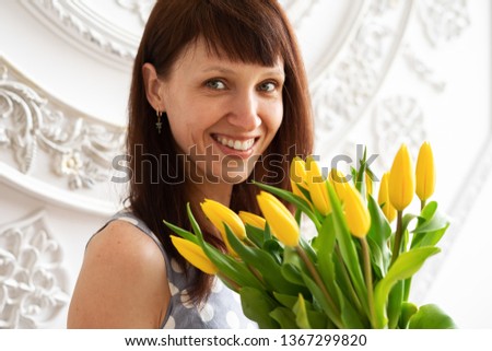 Beautiful brown-haired girl with a large bouquet of yellow tulips against a white wall.