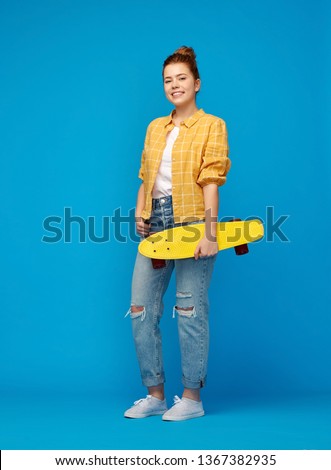 leisure, sport and people concept - smiling red haired teenage girl in checkered shirt and torn jeans with short skateboard over bright blue background