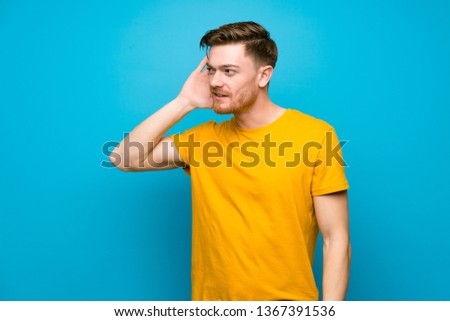 Redhead man over blue wall listening to something by putting hand on the ear
