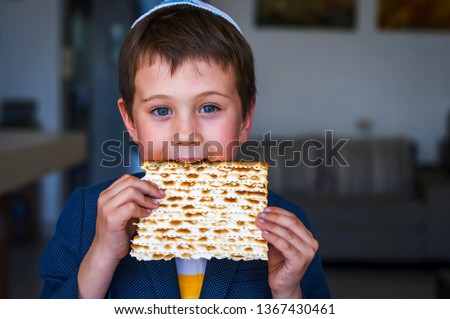 Cute Caucasian Jewish boy holding in his hands and taking a bite from a traditional Jewish matzo unleavened bread. Jewish Passover Pesach concept image. Royalty-Free Stock Photo #1367430461