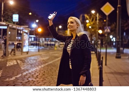 Young blonde woman standing in the city street in the evening, holding smart phone in hand and waiting for taxi or uber. Royalty-Free Stock Photo #1367448803