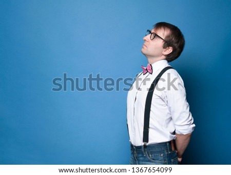 side view of handsome serious man in shirt, suspender, pink bow tie and glasses standing and looking away on blue background with copy space 