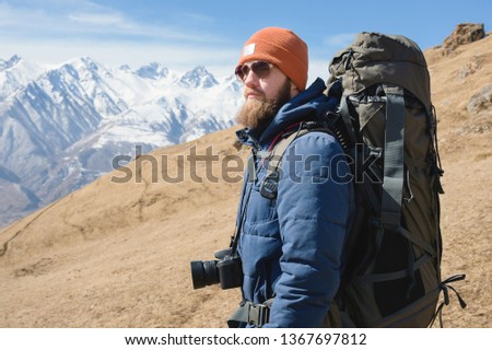 Portrait of a bearded male photographer in sunglasses and a warm jacket with a backpack on his back and a reflex camera on his neck against the background of snow-capped mountains on a sunny day
