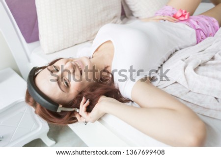 An attractive young brunette with brown hair is lying in her bed in the morning with her head hanging off the edge with headphones and a smartphone and listening to music in a good mood