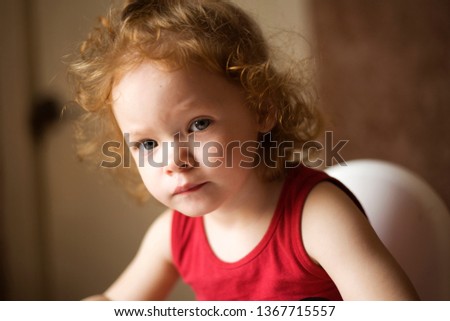 the little red-haired girl is watching cartoons on the phone , a beautiful baby in a red T-shirt is sitting on the highchair and playing games on the phone portrait