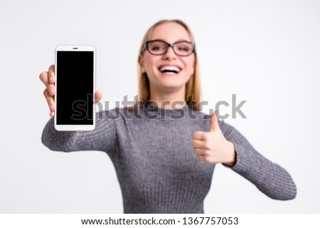 Close up of a mobile cell smart phone with a black screen in hand of young beautiful smart attractive blonde girl in glasses full of emotions smiling with thumb up on white background isolated 
