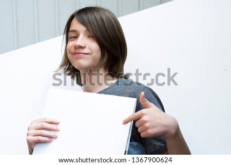 A teenager with long hair recommends smiling, shows a finger on a white blank sheet of paper against a white wall background, on the terrace on a sunny day Photo floor of the body For your advertising