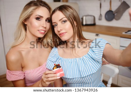 Two charming smiling young woman taking selfie on smartphone, sitting at kitchen, laughing and having fun. Girls party concept