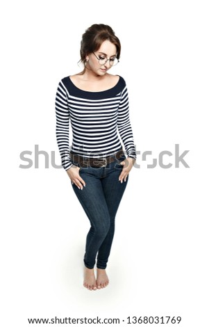 A young woman in a striped tight blouse  and round glasses stands modestly, pressing his hands to his hips. Modestly looks down. Isolated on white background barefoot.