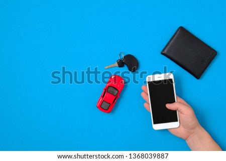 Car rental app concept. Toy car, car key, auto drive license, human hand holding smartphone on blue background. Flat lay composition. Top view.