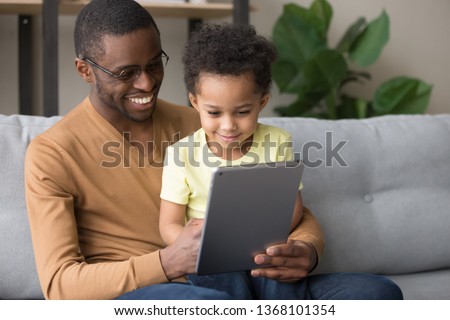 African happy daddy holding son on lap sitting together on couch in room, loving father teach use tablet electronic device, watching cartoon online, play game, spend time at home wireless tech concept