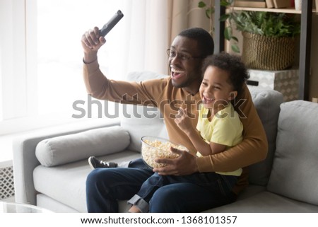 Excited african father raise hand hold remote control celebrate football team victory little son sitting on dad lap family eating popcorn watching sport program on tv, leisure activity at home concept