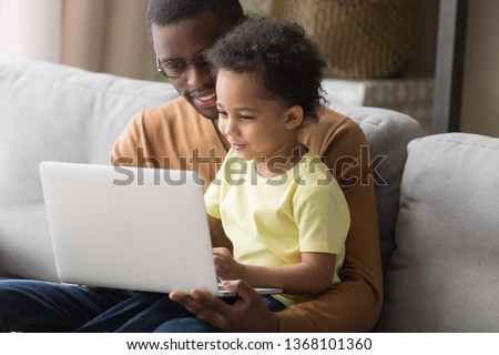 African father holds little son on lap family sitting on sofa at home with notebook, looking at screen watching cartoon educational videos, make video call, having fun enjoy free time together concept Royalty-Free Stock Photo #1368101360
