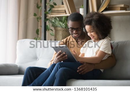 African father holds on lap daughter sitting resting on sofa in living room using digital tablet holding mobile computer watch cartoons educational online program, buying shopping via internet concept Royalty-Free Stock Photo #1368101435