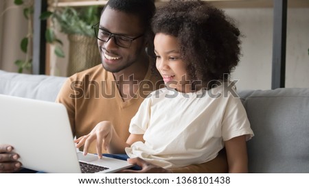 Biracial daughter sitting on dad lap, family spends day use computer, father teach kid girl typing on laptop, surfing internet, chatting online with friend. Modern tech usage, leisure at home concept