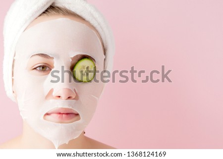 Beautiful young woman is applying a cosmetic tissue mask on a face with cucumber on a pink background. Healthcare and beauty treatment and technology concept.