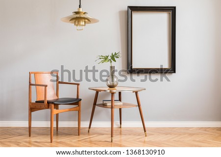 Stylish and bright retro interior with design chair , gold lamp and small table with vase. Black mock up frame on the gray background wall. Minimalistic concept of sitting room. Real photo. 