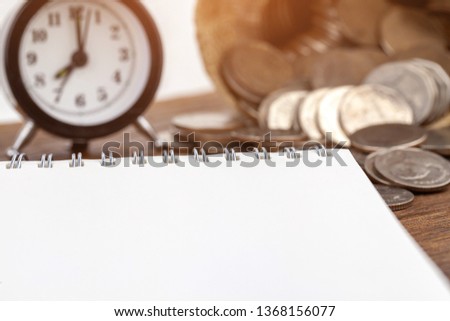 Blank notepad, alarm clock and coins on it on office wooden table
