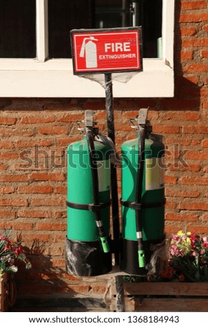 Two green fire extinguisher with the red sign and white text and symbolic icon on brick wall and window background.