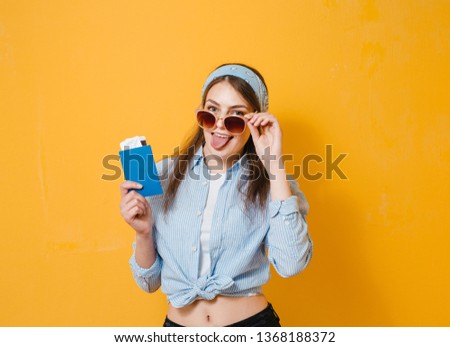 A portrait of a beautiful  girl with sunglasses posing with gestures and holding a passport with a ticket over yellow background