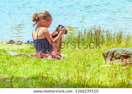 Little girl taking pictures sitting near the river on the grass.Cute little girl sitting on the grass on sunny summer day and taking picture with camera. Summer holidays.