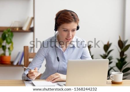 Businesswoman wearing headphones watching video webinar making conference online call writing notes talking, focused woman study online looking at laptop listening translating lecture course