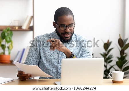 Focused african american businessman working with laptop documents in office holding papers preparing report analyzing work results, black male analyst doing paperwork at workplace using computer Royalty-Free Stock Photo #1368244274
