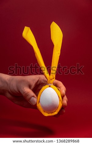 The hand that holds the egg in a yellow scarf, the fingers hold the egg, the egg is on the hand, the composition of the hand and the egg in a yellow scarf on a burgundy background