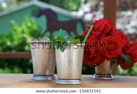 Mint juleps in traditional silver cups with red roses and horse shape in background Royalty-Free Stock Photo #136831541
