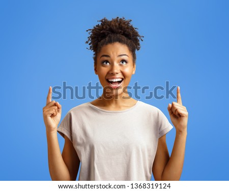 Amazed girl looks up, indicates with two forefingers upwards at copy space, shows something. Photo of african american girl on blue background. Emotions and pleasant feelings concept. Royalty-Free Stock Photo #1368319124
