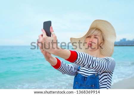 beautiful young blonde caucasian woman on vacation in straw hat, striped blouse nad denim overalls making selfie on smartphone by the amazing blue sea background