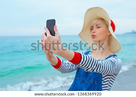 beautiful young blonde caucasian woman on vacation in straw hat, striped blouse nad denim overalls making selfie on smartphone by the amazing blue sea background