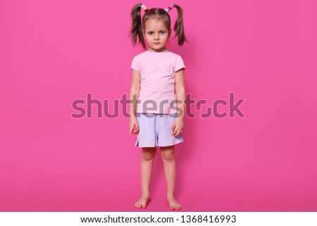 Tired magnetic small girl with pigtails is focused on camera, looks serious, wearing light pink t shirt and casual shorts, having colourful scrunchies in her hair. Childhood and emotions concept.