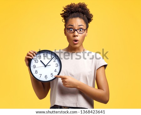 Time to work! Student with shocked face holds clock and points to them. Photo of african american girl wears casual outfit on yellow background. Emotions and pleasant feelings concept. Royalty-Free Stock Photo #1368423641