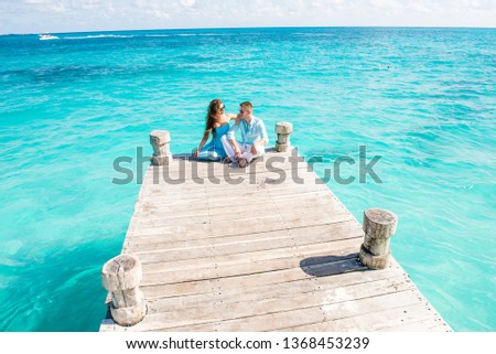 Young beautiful couple in blue and white clothes are sitting at the wooden pier. Drone view. Woman put hand on guy's shoulder and tell him something. Turquoise color water behind them. Couple vacation Royalty-Free Stock Photo #1368453239