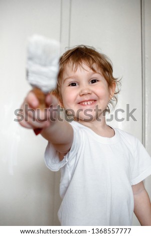 A little naughty girl in a white t-shirt paints the wall with white paint. reaching into the camera with a brush