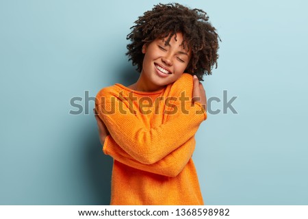 Charming gorgeous Afro woman keeps eyes closed, smiles with pleasure, shows white teeth feels comfort, hugs herself, wears orange jumper, tilts head, models over blue background, has high self esteem Royalty-Free Stock Photo #1368598982