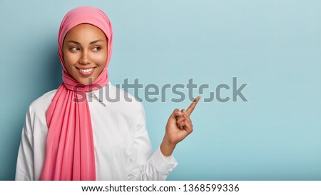 Smiling black woman has cheerful expression, points away with fore finger, shows blank space on right corner, has modest look, wrapped in pink veil, isolated over blue background. Look at this Royalty-Free Stock Photo #1368599336