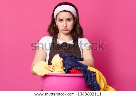 Studio shot of brunette housekeeper with pouty lips, has upset and tired facial expression, wears brown apron and casual t shirt, poses against pink background, wants to have rest, looks exuasted.
